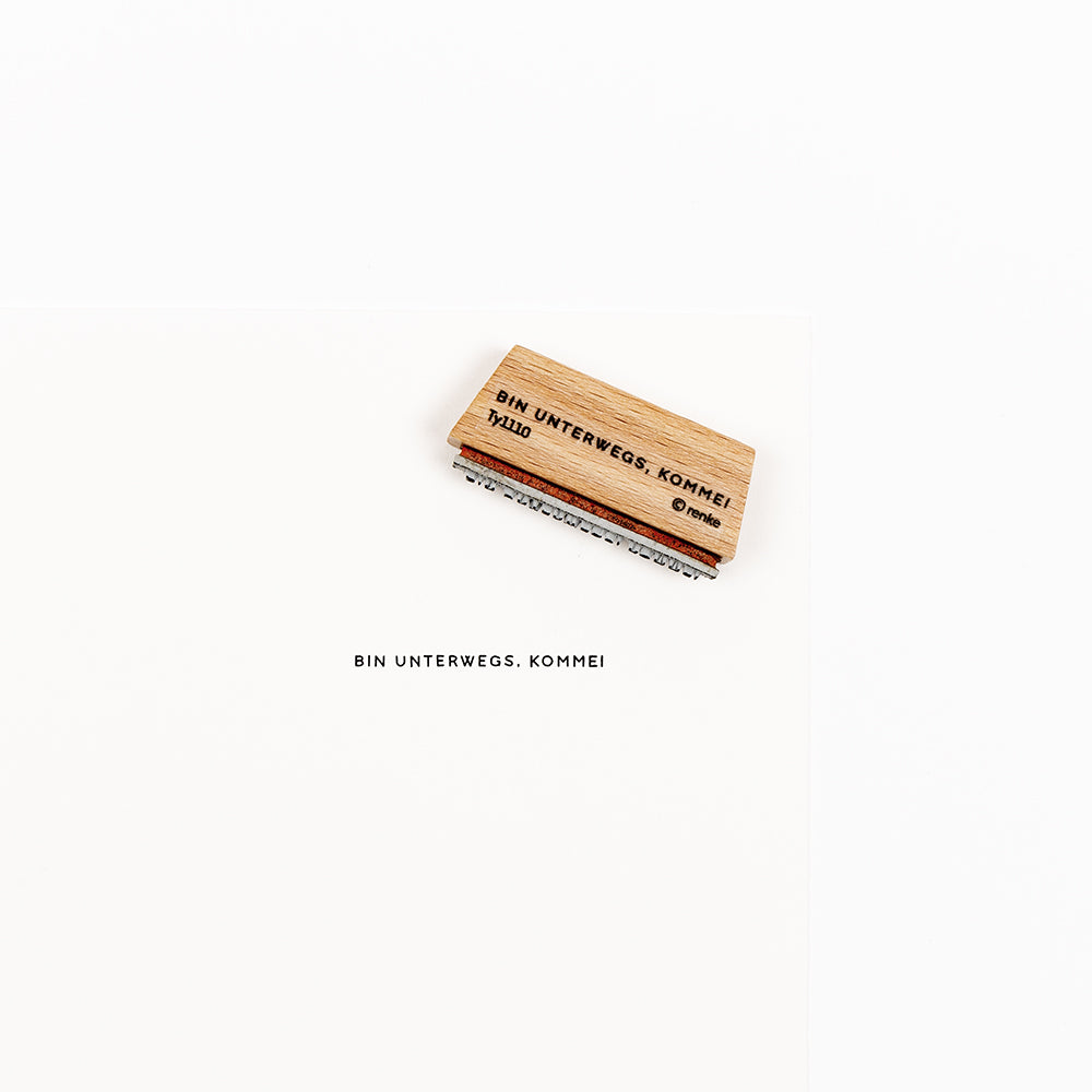 Ein Mini-Typo-Stempel aus Holz mit der Aufschrift BIN UNTERWEGS, KOMME!, aufgedruckt auf weißem Papier.
