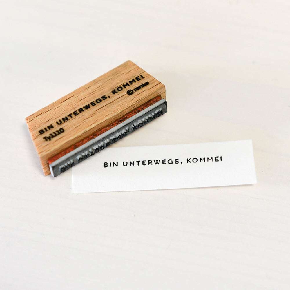Ein Holzstempel und gestempeltes Papier von Alexandra Renke mit "BIN UNTERWEGS, KOMME!" auf weißem Untergrund - eine tolle Inspiration für deine Papierprojekte oder den kreativen Einsatz von Stempelkissen.