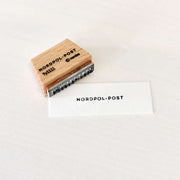 Ein Holzstempel von Alexandra Renke mit dem Schriftzug Nordpol-Post ruht neben gestempeltem Text auf einer weißen Karte und zeigt ein reizvolles Stempelmotiv.