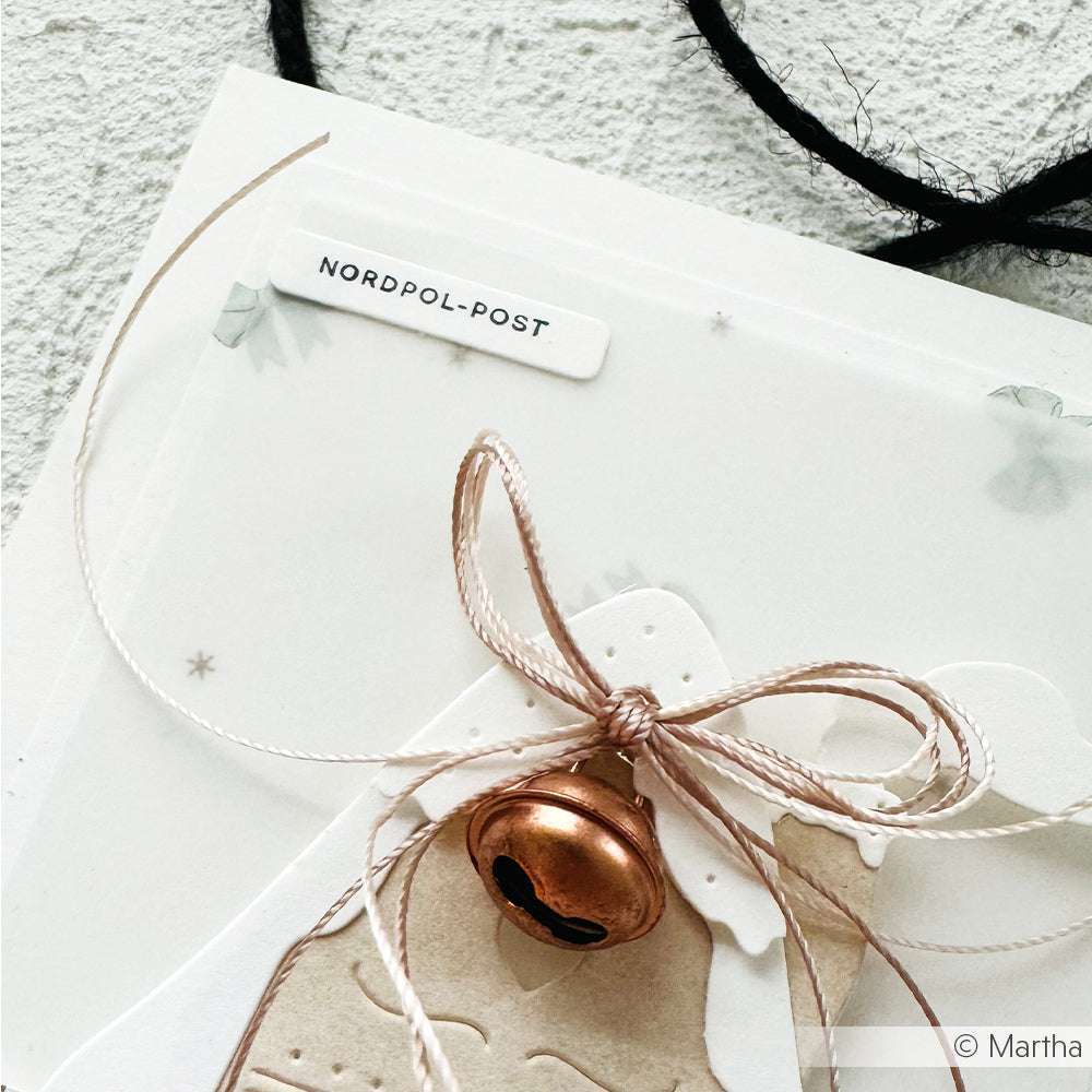 Eine Weihnachtskarte von Alexandra Renke mit einem Metallglöckchen und einer Schleife, einem Nordpol-Post-Etikett und einem weißen, strukturierten Hintergrund - ideal zum Kombinieren mit Ihren Lieblings-Holzstempeln für festliche Bastelarbeiten.