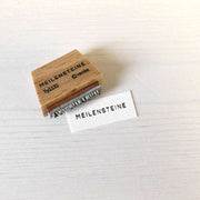 Ein Holzstempel von Alexandra Renke mit der Aufschrift MEILENSTEINE ruht neben seinem Aufdruck auf weißem Papier - ideal, um Meilensteine in Ihren Papierprojekten zu markieren.