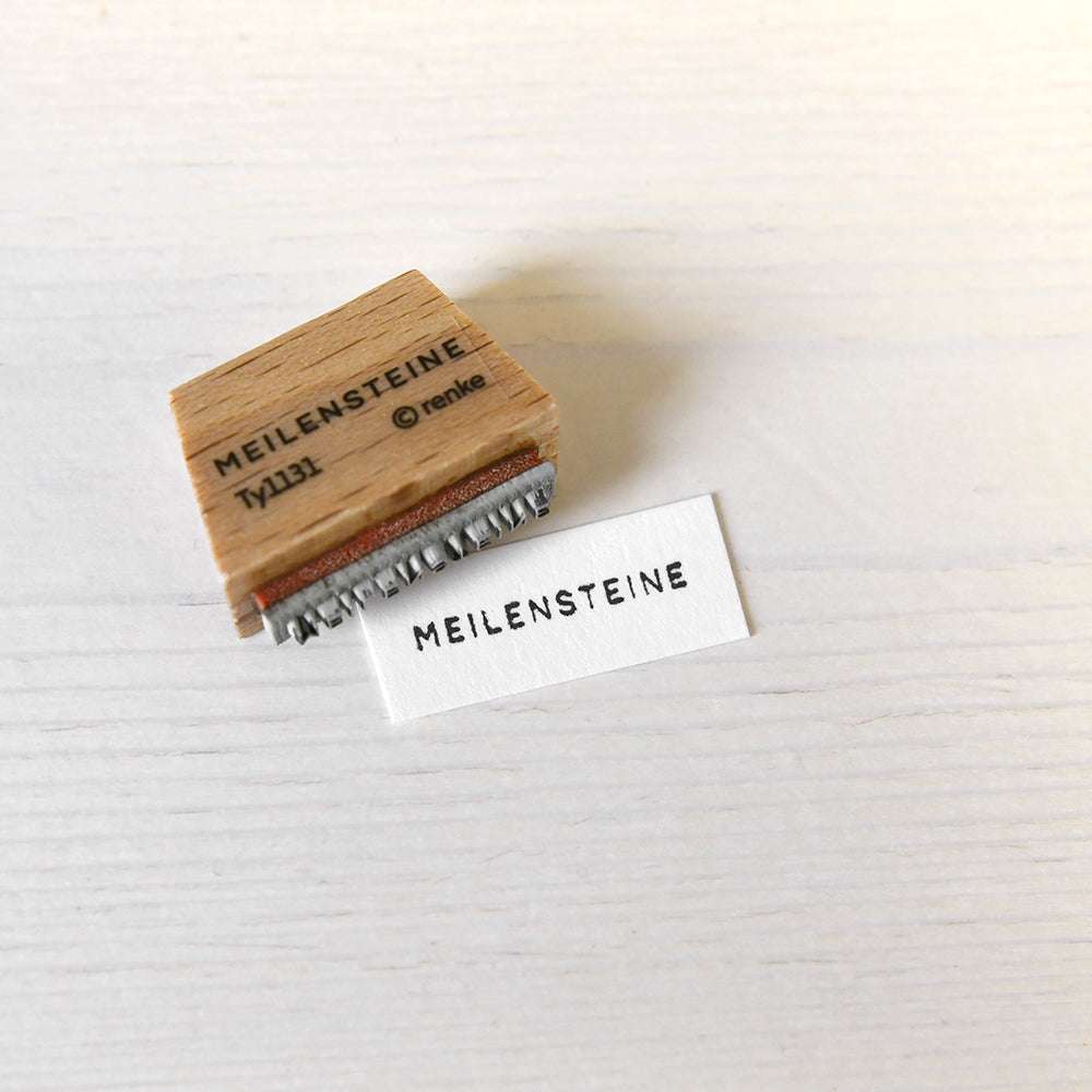 Ein Holzstempel von Alexandra Renke mit der Aufschrift MEILENSTEINE ruht neben seinem Aufdruck auf weißem Papier - ideal, um Meilensteine in Ihren Papierprojekten zu markieren.