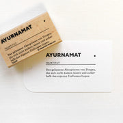 Ein brauner Holzstempel von Alexandra Renke aus hochwertigem Holz und eine gestempelte Karte mit dem Motiv "AYURNAMAT" sowie einer deutschen Definition.