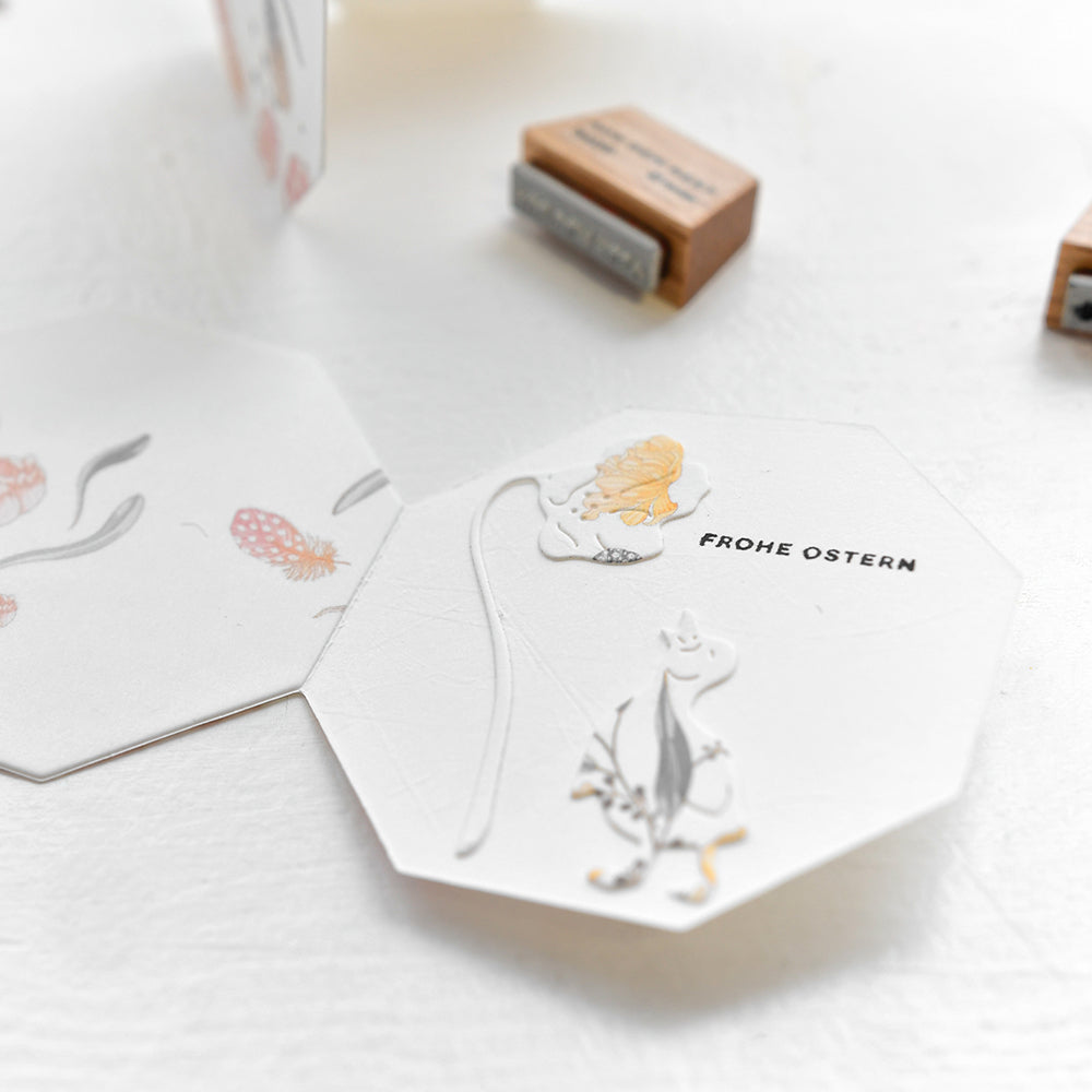 Sechseckige Karte von Alexandra Renke mit floralen Illustrationen und "Frohe Ostern"-Text auf weißer Fläche, abgebildet mit einem Holzstempel und Stempelkissen in der Nähe.