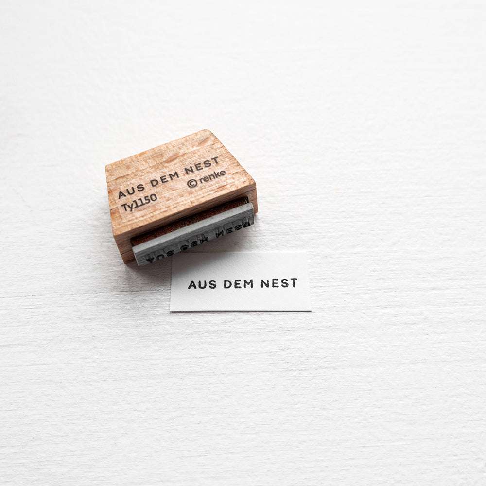Ein Alexandra Renke Holzstempel mit der Aufschrift AUS DEM NEST steht neben einem gestempelten Text auf weißem Papier - ideal für kreative Papierprojekte.
