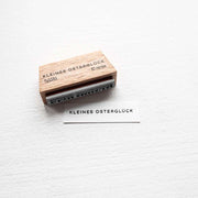 Ein Holzstempel von Alexandra Renke stempelt "KLEINES OSTERTGLÜCK" auf weißes Papier - ideal, um Ihren Osterglück-Basteleien Mini-Typo-Flair zu verleihen.