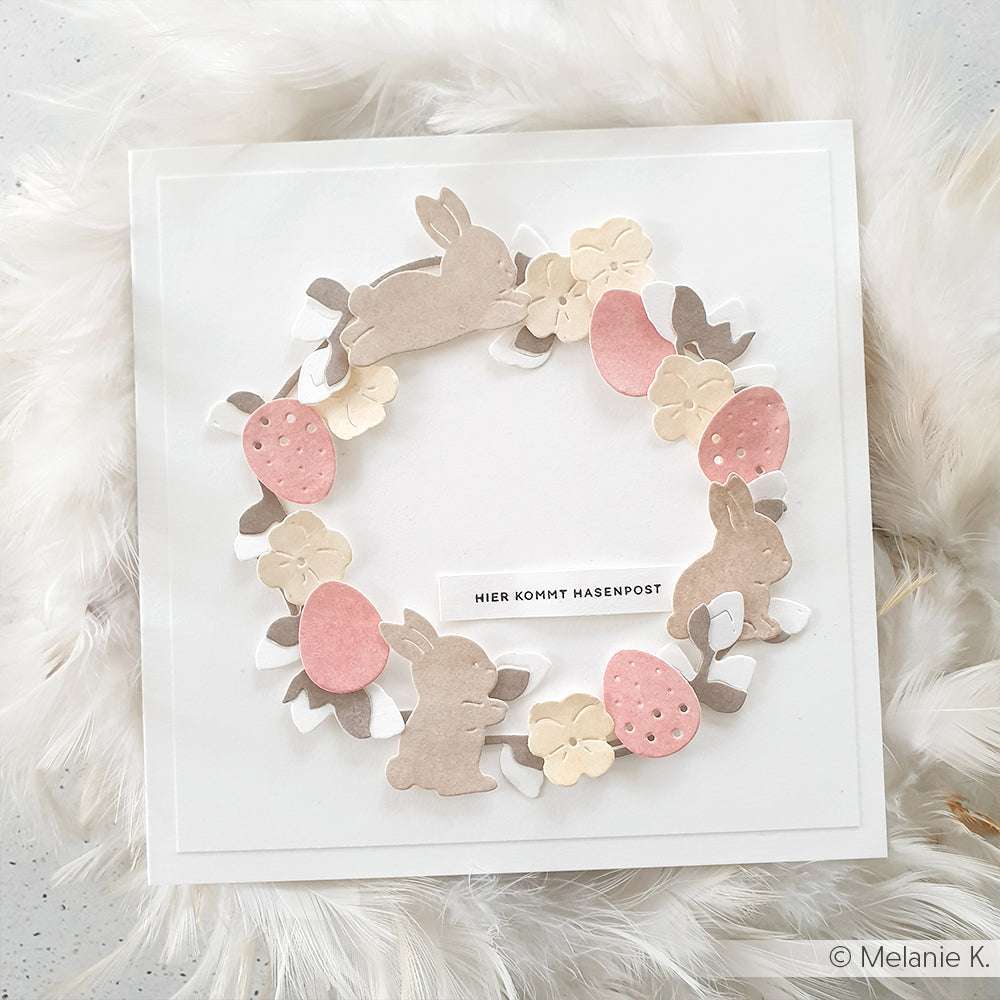 Eine quadratische Karte von Alexandra Renke mit einem Kranz aus Papierhasen, Eiern und Blumen, gestempelt mit Holzstempel und Stempelkissen, mit einem deutschen Text in der Mitte.