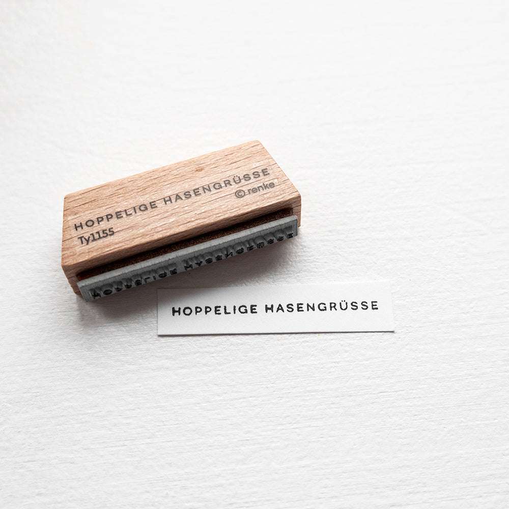 Ein Holzstempel von Alexandra Renke und ein Papier mit der Aufschrift HOPPELIGE HASENGRÜSSE in schwarzen Buchstaben - ideal für kreative Papierprojekte.