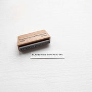 Ein Holzstempel von Alexandra Renke und ein Zettel mit der Aufschrift "FLAUSCHIGE OSTERGRÜSSE" in der Schriftart Mini Typo sind auf einem weißen Hintergrund abgebildet.
