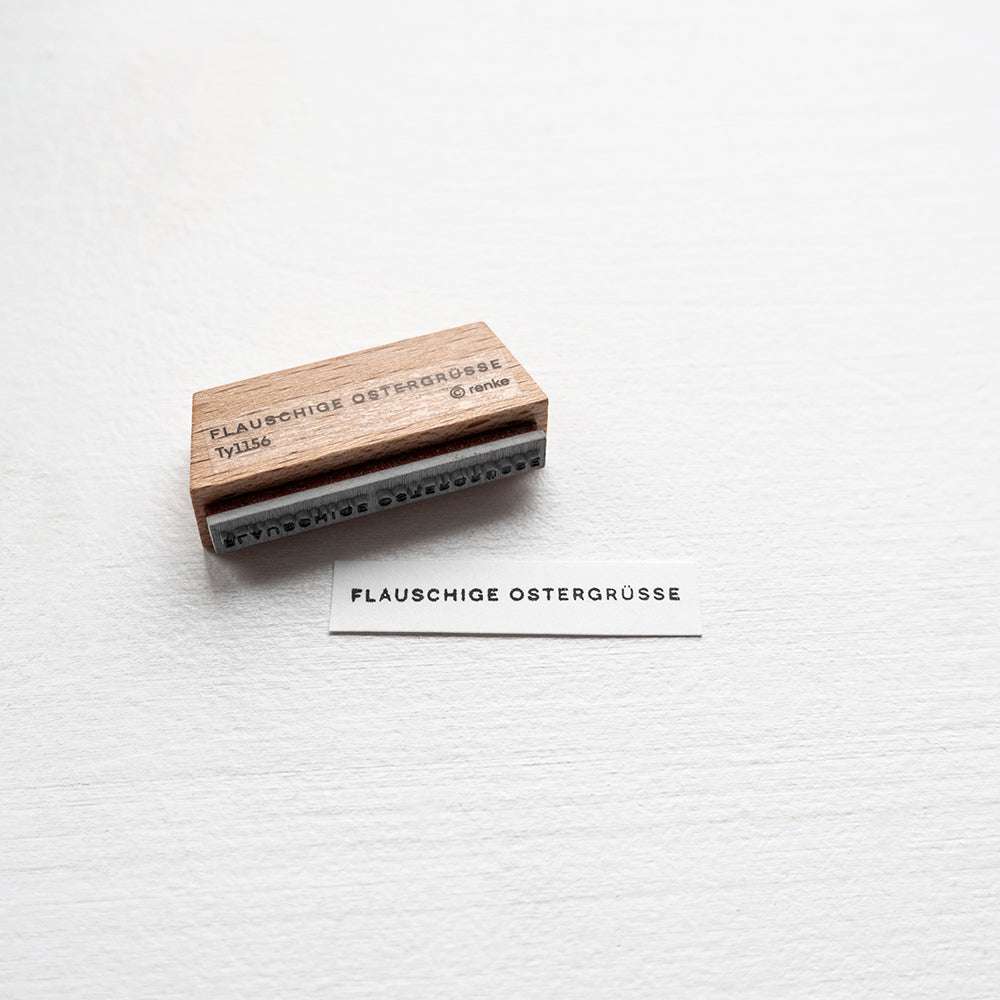 Ein Holzstempel von Alexandra Renke und ein Zettel mit der Aufschrift "FLAUSCHIGE OSTERGRÜSSE" in der Schriftart Mini Typo sind auf einem weißen Hintergrund abgebildet.