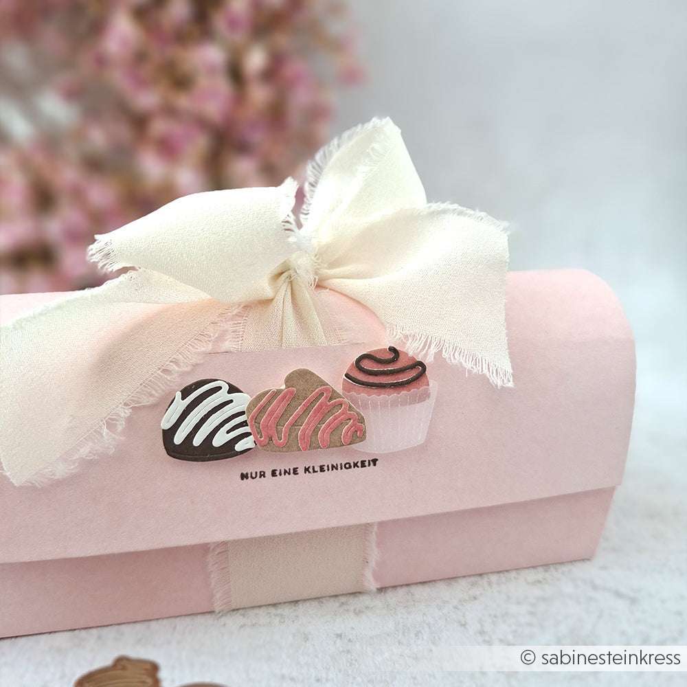 Eine rosafarbene Alexandra Renke Geschenkbox mit cremefarbener Schleife und Schokoladentrüffel-Illustrationen, ideal für Papierprojekte oder Stempelgummi-Design-Inspiration, ausgestellt auf einer hellen Oberfläche.