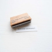 Der Alexandra Renke Holzstempel und gestempeltes Papier zeigen "NUR EINE KLEINIGKEIT" auf Weiß - ideal für kreative Papierprojekte mit deinem Lieblings-Stempelkissen.