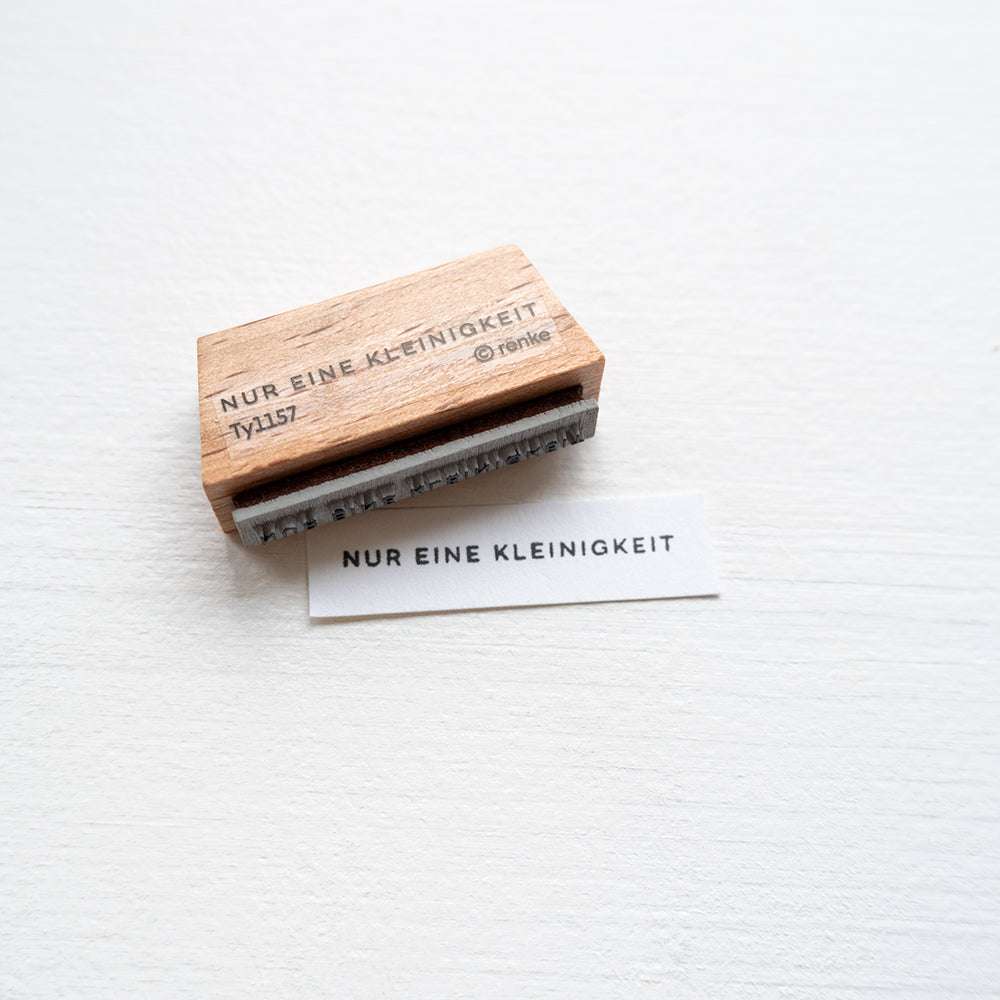 Der Alexandra Renke Holzstempel und gestempeltes Papier zeigen "NUR EINE KLEINIGKEIT" auf Weiß - ideal für kreative Papierprojekte mit deinem Lieblings-Stempelkissen.