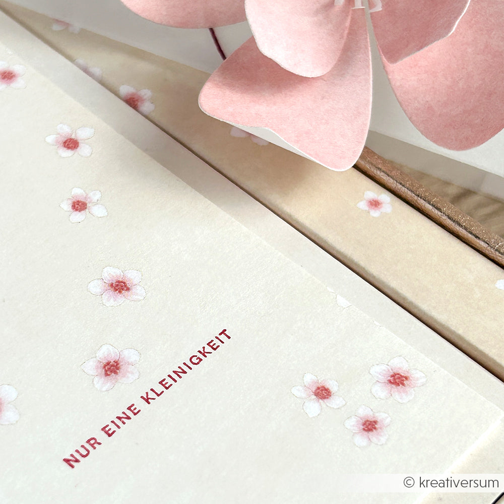 Weiße Karte von Alexandra Renke mit kleinen rosa Blumen, gestempelt mit einem Holzstempel und dem deutschen Text "NUR EINE KLEINIGKEIT" neben einer rosa Papierblume.