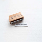 Ein Holzstempel von Alexandra Renke und gestempeltes Papier, beides mit "HIPP HIPP HURRA" auf weißer Oberfläche - ideal für kreative Papierprojekte mit einzigartigen Stempelgummi-Designs.