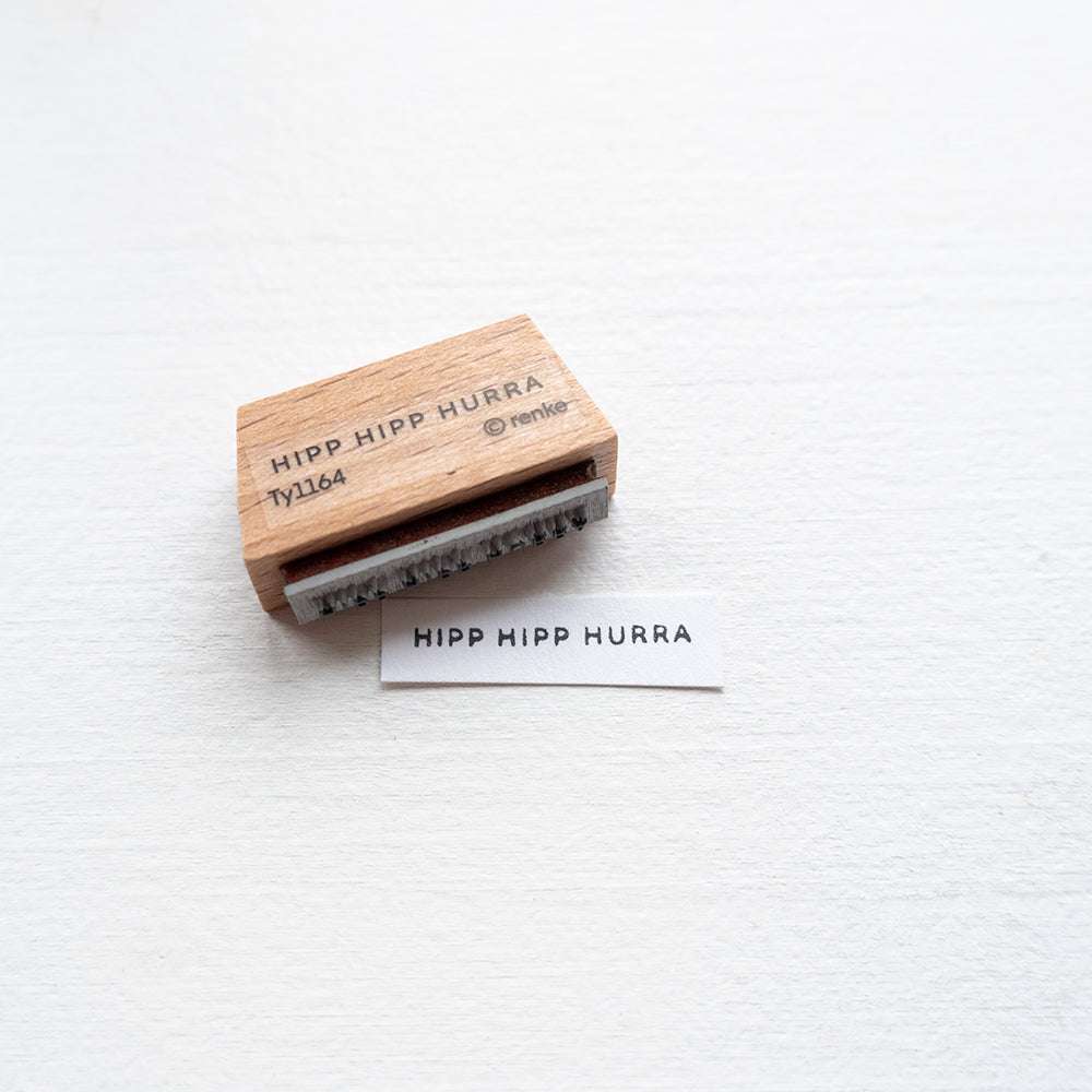 Ein Holzstempel von Alexandra Renke und gestempeltes Papier, beides mit "HIPP HIPP HURRA" auf weißer Oberfläche - ideal für kreative Papierprojekte mit einzigartigen Stempelgummi-Designs.
