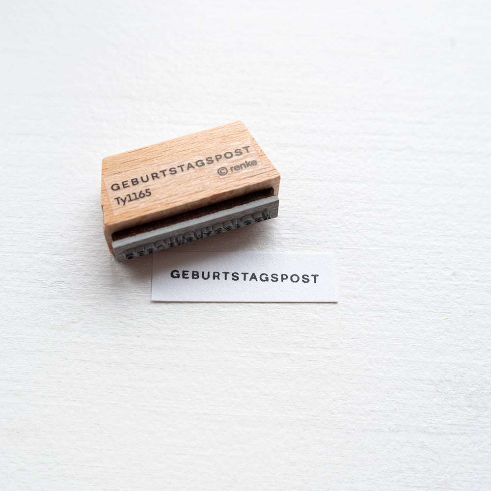 Der Alexandra Renke Holzstempel mit der Aufschrift Geburtstagspost ist neben gestanztem Text auf weißem Papier abgebildet, ideal für kreative Papierprojekte auf hellem Untergrund.