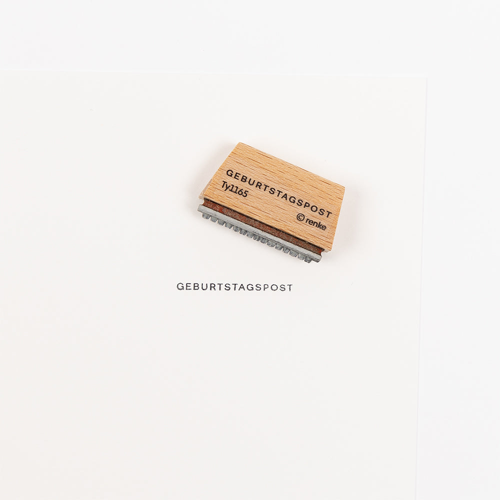 Oben ist ein Holzstempel mit der Aufschrift „GEBURTSTAGSPOST“ (Holzstempel) zu sehen, der auf weißem Papier den gleichlautenden Text abgedruckt hat.