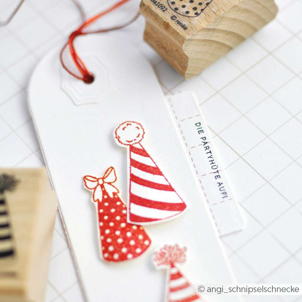 Ein weißer Anhänger von Alexandra Renke, verziert mit roten Partyhüten, einer Schnur und einem kleinen Etikett auf einem Gitter - perfekt für Papierprojekte oder in Kombination mit Ihren Lieblings-Holzstempeln.