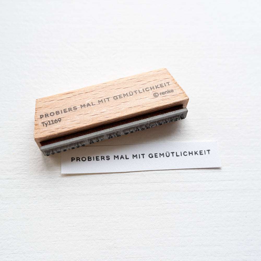 Ein hochwertiger Holzstempel von Alexandra Renke und gestempeltes Papier mit "PROBIERS MAL MIT GEMÜTLICHKEIT" auf weißem Untergrund - geeignet für Stempelkissen.