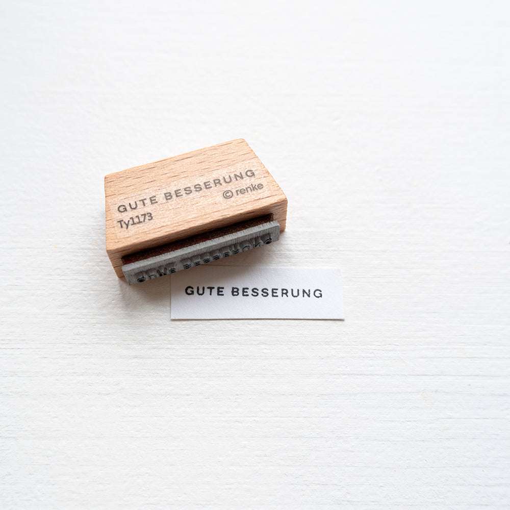 Ein Holzstempel der Marke Alexandra Renke und ein Papier mit dem Aufdruck Gute Besserung liegen auf einer weißen Oberfläche.