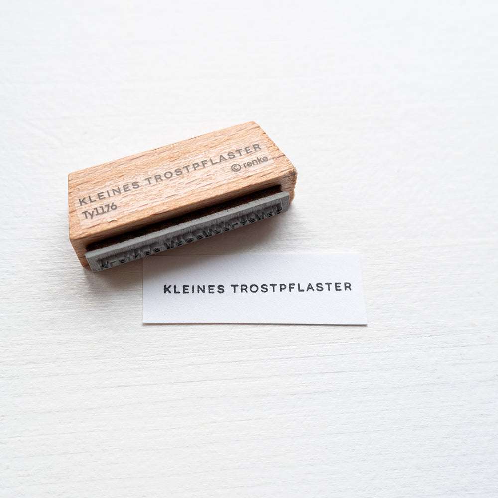 Ein Holzstempel von Alexandra Renke mit Stempelgummi zeigt den Text KLEINES TROSTPFLASTER, das Motiv ist auf Papier gedruckt.