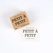 Ein hölzerner Alexandra Renke Holzstempel und ein gestempeltes Papier mit "PETIT À PETIT, LITTLE BY LITTLE" in schwarz-ideal für detailreiche Motive auf Ihren Papierprojekten.