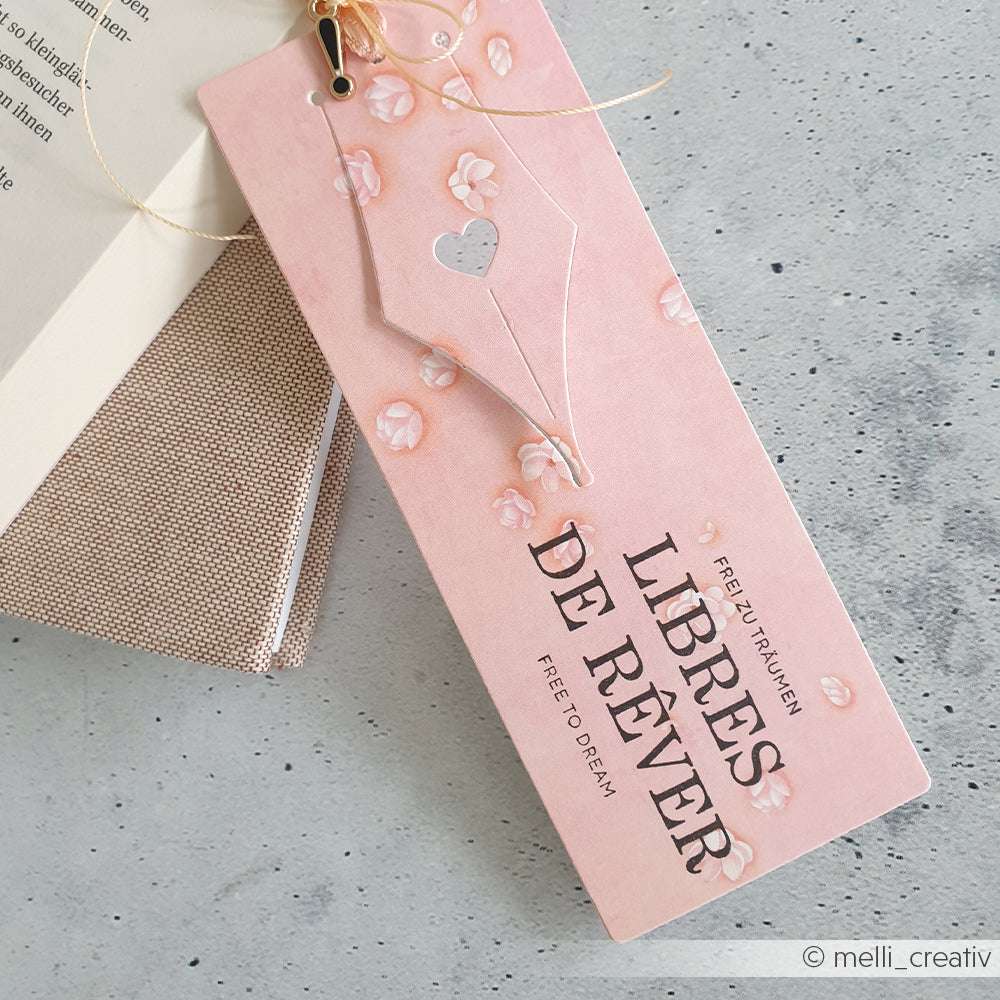 Ein rosa geblümtes Lesezeichen von Alexandra Renke aus hochwertigem Holz mit der Aufschrift "LIBRES DE RÊVER, Free to Dream" liegt neben einem Buch.