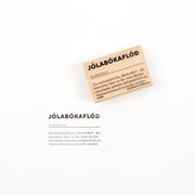 Ein Holzstempel und sein Stempelabdruck, auf denen jeweils das isländische Wort „Jólabókaflóð“ und dessen Definition zu sehen sind.