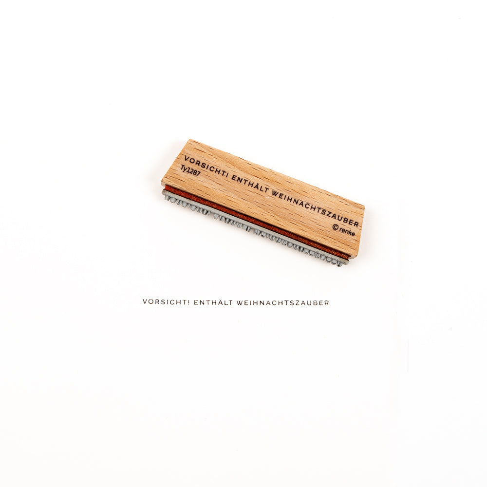 Ein Stempel aus Holz mit der Aufschrift „VORSICHT! ENTHÄLT WEIHNACHTSZAUBER“, der neben dem Stempel selbst ausgestellt ist.