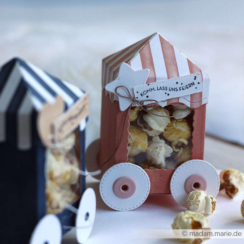 Gestreifte Zirkuswagen-Papier-Popcornboxen von Alexandra Renke aus hochwertigem Material, gefüllt mit Popcorn und mit einem "Komm, lass uns feiern!"-Schild versehen.
