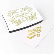 Ein weißes Tsukineko VersaFine Stempelkissen liegt neben einem Anhänger, der mit Pigment-Tinten mit einem goldenen Blumenmuster gestempelt wurde.