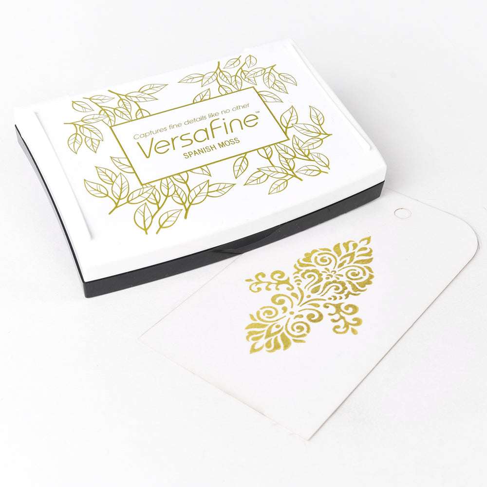 Ein weißes Tsukineko VersaFine Stempelkissen liegt neben einem Anhänger, der mit Pigment-Tinten mit einem goldenen Blumenmuster gestempelt wurde.