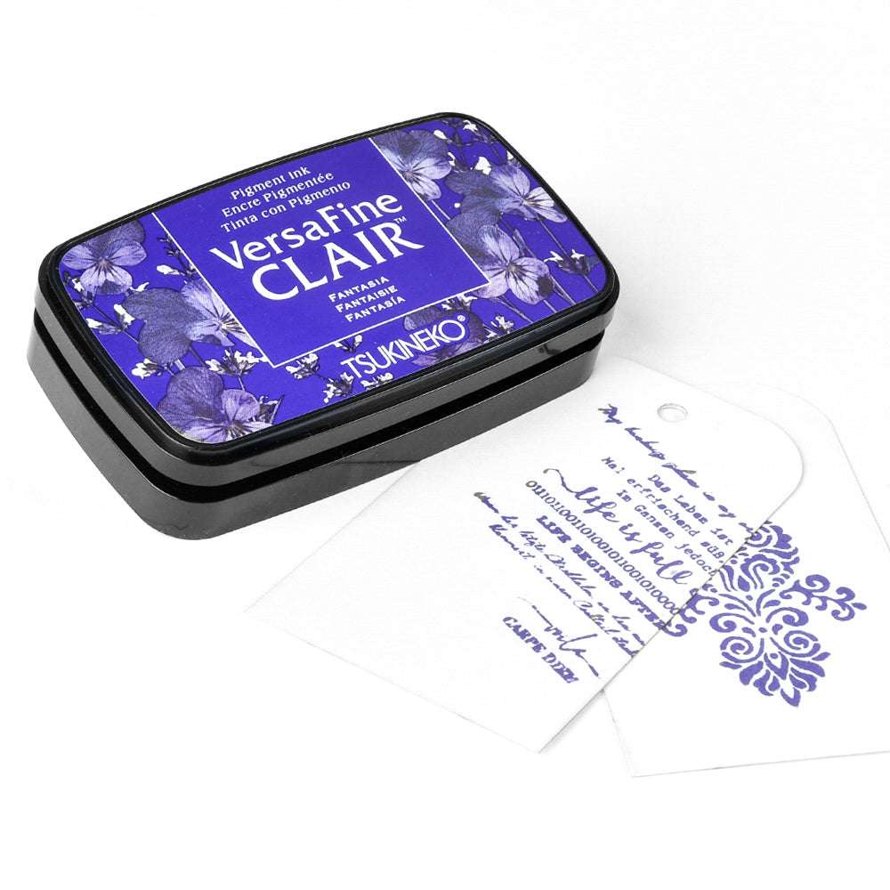 Tsukineko VersaFine Clair Stempelkissen mit blauem Blumenetikett, daneben zwei weiße Anhänger mit Designs in blauer Pigment-Tinte gestempelt.