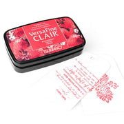 Ein rotes Tsukineko VersaFine Clair Stempelkissen und zwei weiße Anhänger, die mit roten Tsukineko-Tintenmustern und Text bestempelt sind.