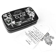 Ein schwarzes Tsukineko VersaFine Clair Stempelkissen liegt neben einem weißen Anhänger mit schwarzer Schrift und einem schablonierten Blumenmuster.