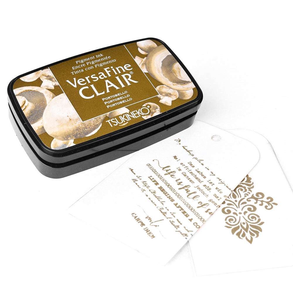 Das Tsukineko VersaFine Clair Stempelkissen in Portobello liegt neben zwei weißen Anhängern, die mit VersaFine-Tinte bestempelt und mit Text sowie Mustern verziert sind.