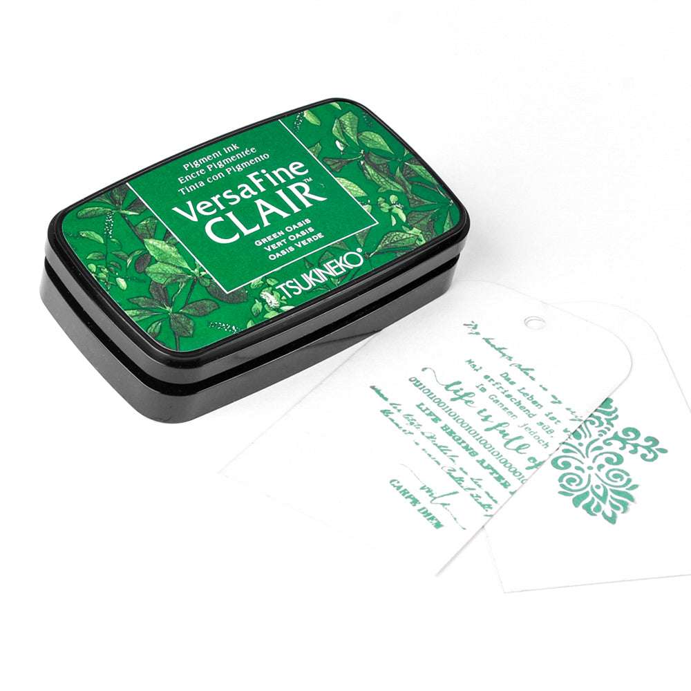 Tsukineko VersaFine CLAIR grünes Stempelkissen mit zwei weißen, grün gestempelten Anhängern, die Text und dekorative Motive zeigen.