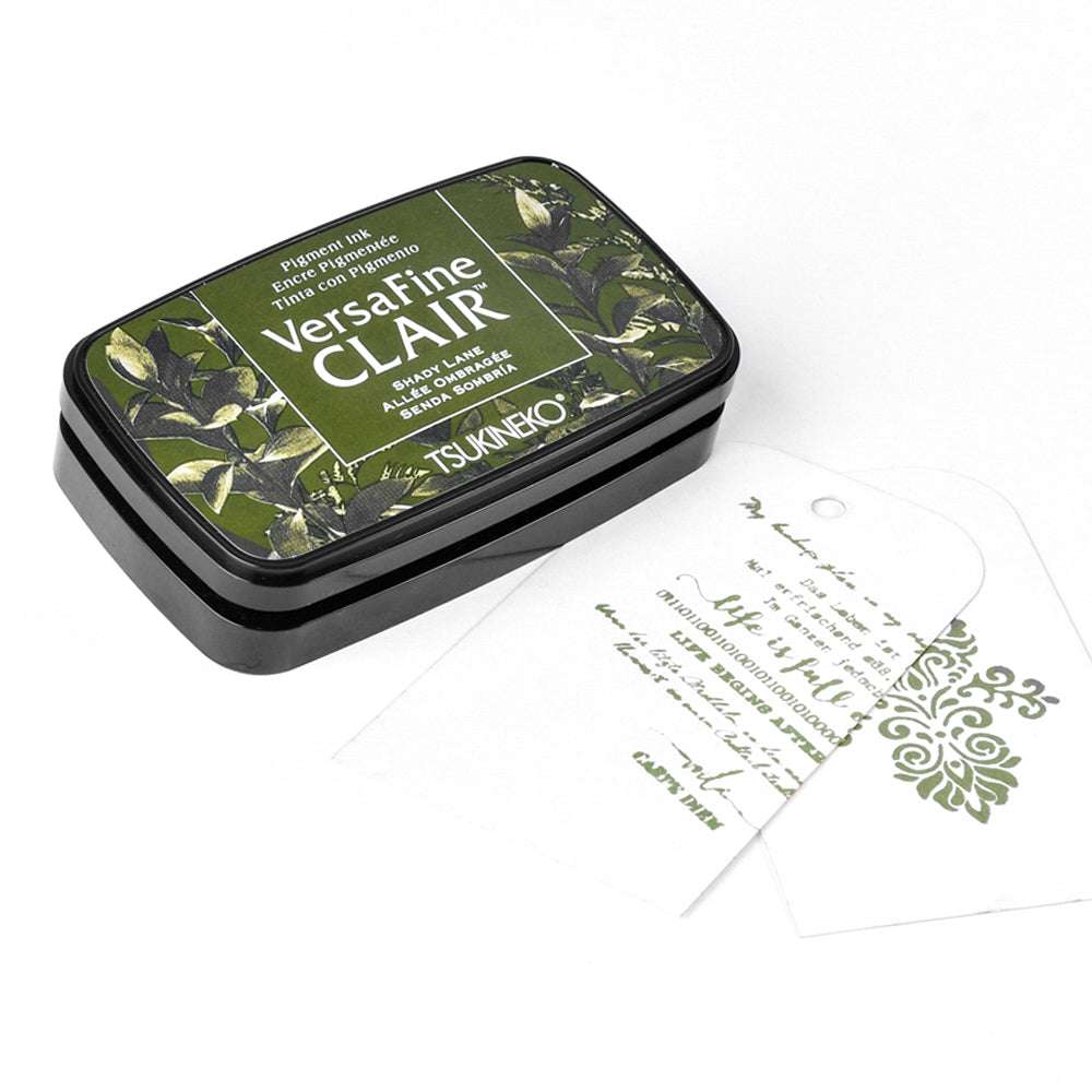 Ein grünes Tsukineko Versafine Clair Stempelkissen liegt neben zwei weißen Anhängern mit grünen Stempelmotiven und dem Text "Pigment-Tinten".