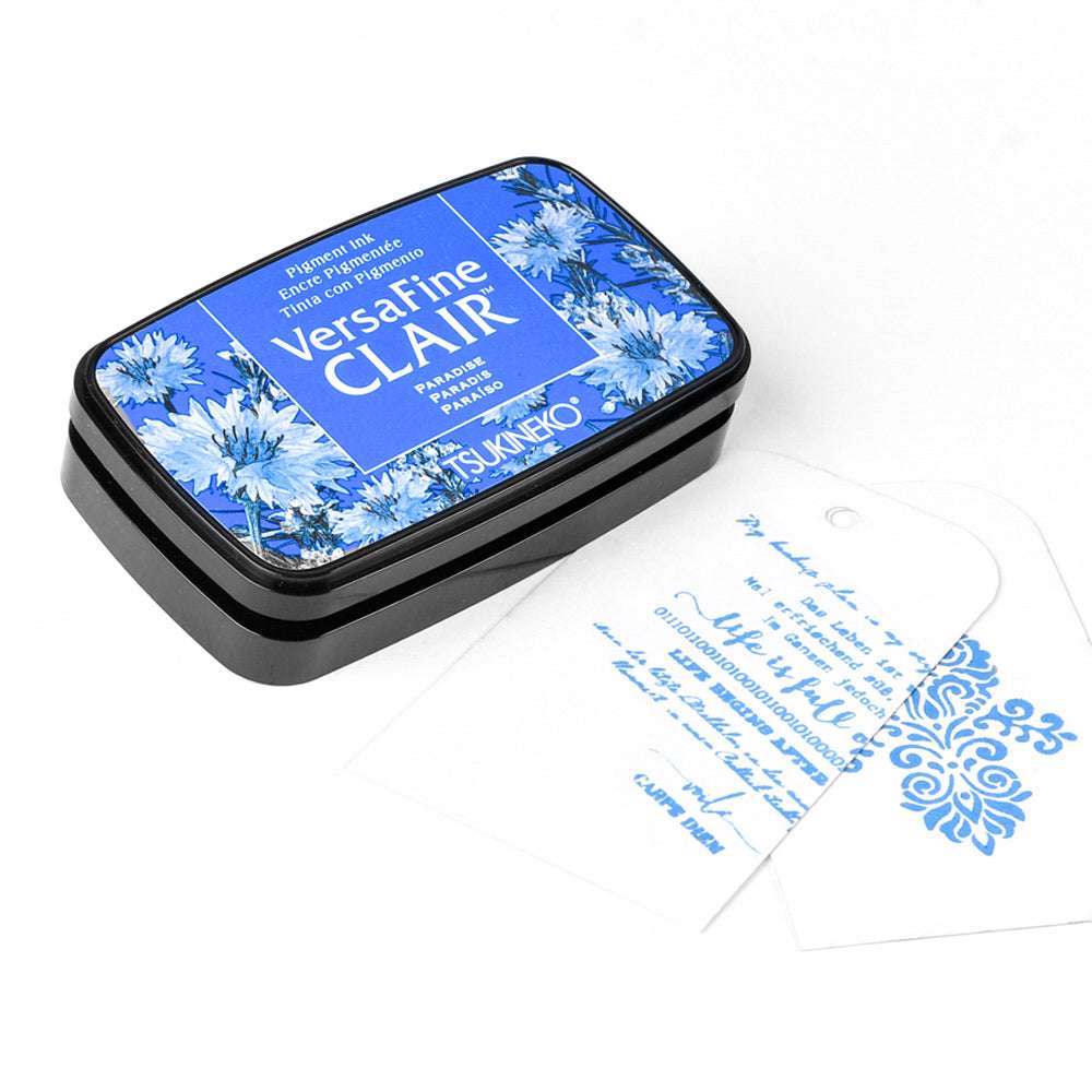 Tsukineko VersaFine Clair blaues Stempelkissen mit Blumenmuster neben zwei weißen Anhängern, die jeweils mit kräftigen blauen Pigmenttintenmustern gestempelt sind.