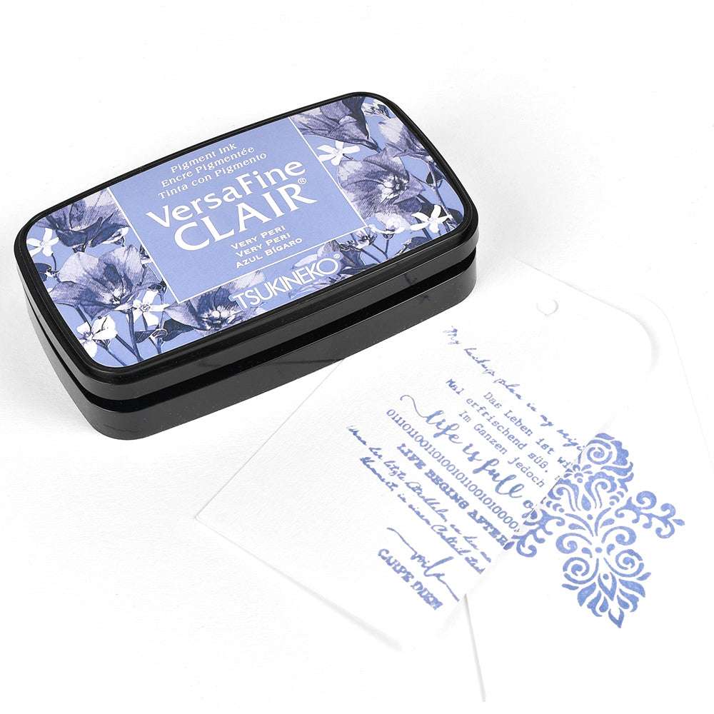 Ein blaues Tsukineko Versafine Clair Stempelkissen liegt neben einem weißen Anhänger, der mit blauem Ziertext und Mustern bestempelt ist.
