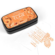 Ein orangefarbenes Stempelkissen Tsukineko VersaFine Clair liegt neben einem weißen Anhänger, der mit orangefarbenem Ziertext gestempelt ist.