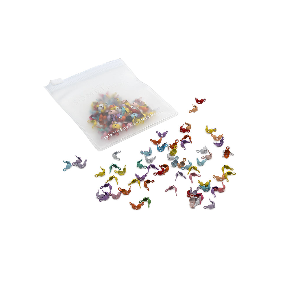 Aus einem kleinen durchsichtigen Beutel quellen bunte, winzige, geschwungene Plastikfiguren in verschiedenen Formen und Farben hervor, die an Crimp-Spitzen und Quetschkalotten erinnern, wie sie häufig in der Schmuckherstellung verwendet werden.