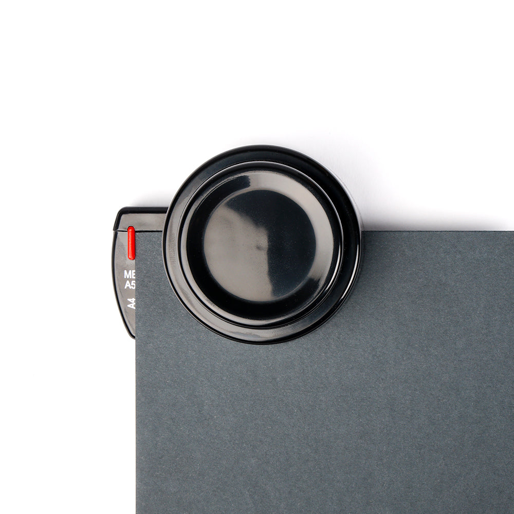 Ein schwarzes Vergrößerungsglas ruht auf einem dunkelgrauen Alexandra Renke Notizbuch mit rotem Reiter - ideal für Ihr nächstes Buchbindeprojekt oder zum Löcher stanzen mit einem Stanzer für Bindescheiben.