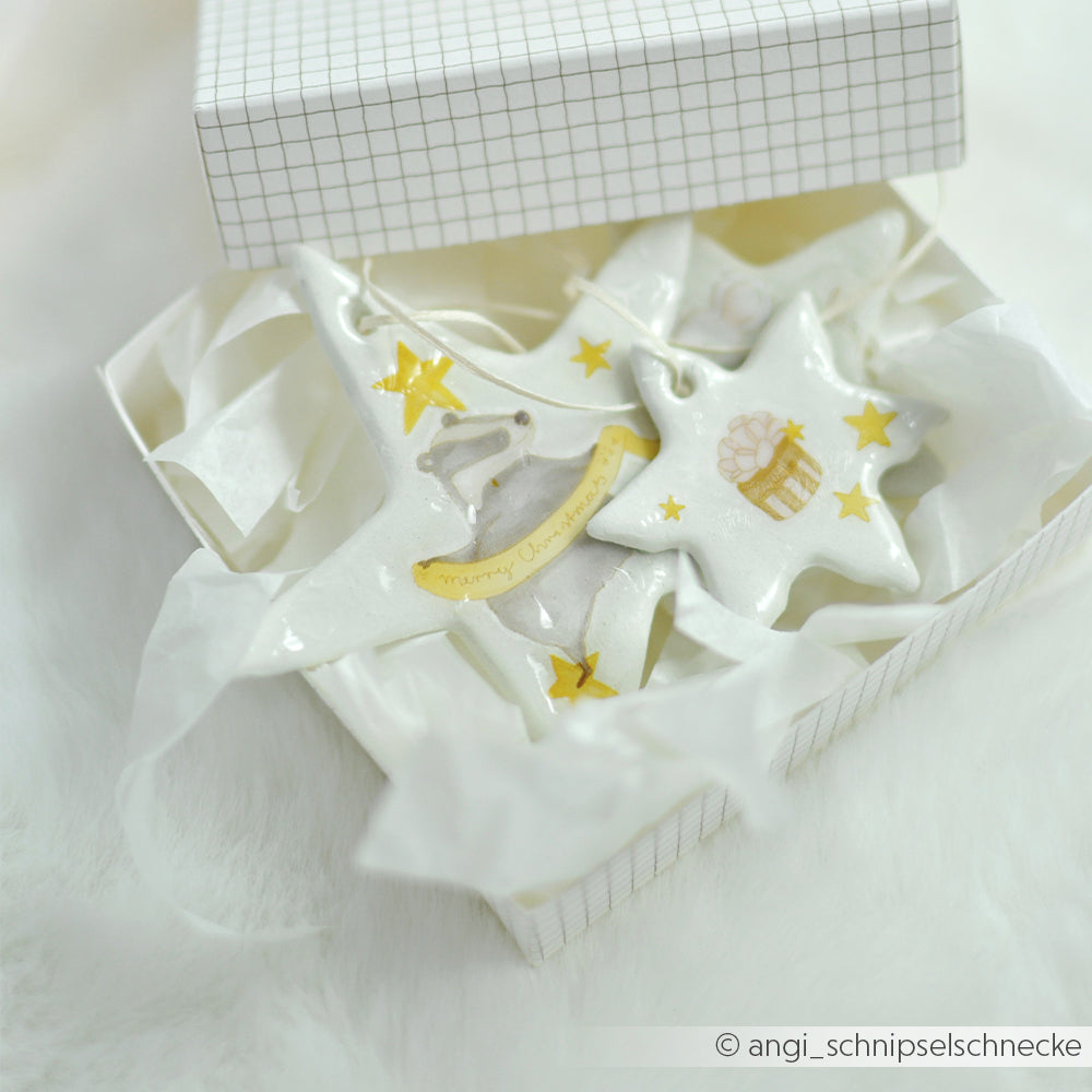 Zwei sternförmige Ornamente aus weißem, lufttrocknendem Ton in einer Geschenkbox, eines davon mit einem Dachs.