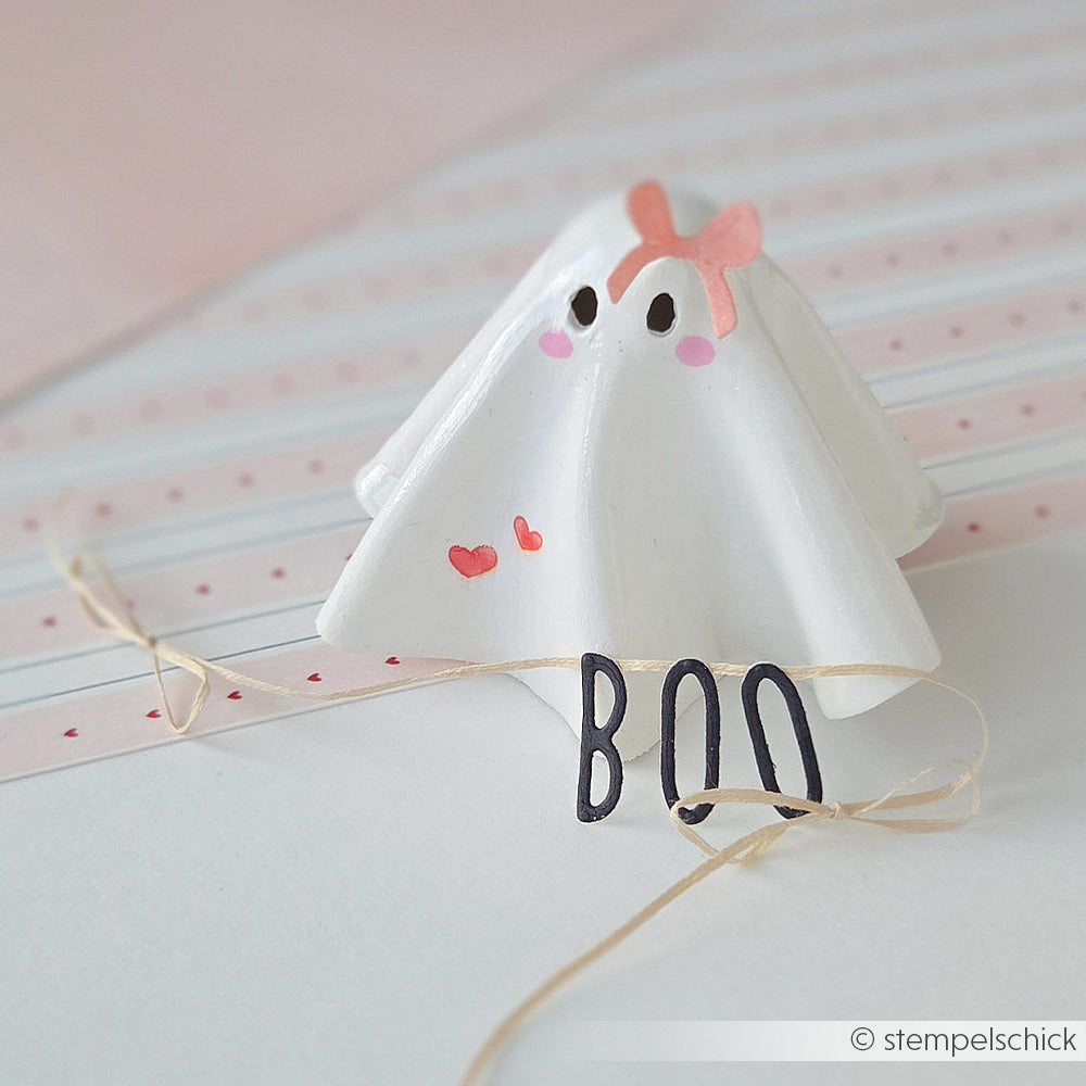 Eine kleine Geisterfigur aus weißer Modelliermasse mit einer rosa Schleife und der Aufschrift BOO auf der Vorderseite.