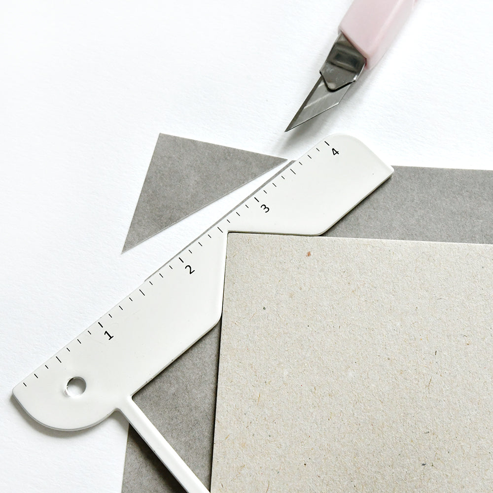 Ein Vaessen-Lineal, ein Bastelmesser und graue Papierbögen, die auf weißem Papier angeordnet sind - ideale Werkzeuge für Buchprojekte oder die Erstellung von individuellen Buchhüllen-Notizbüchern.