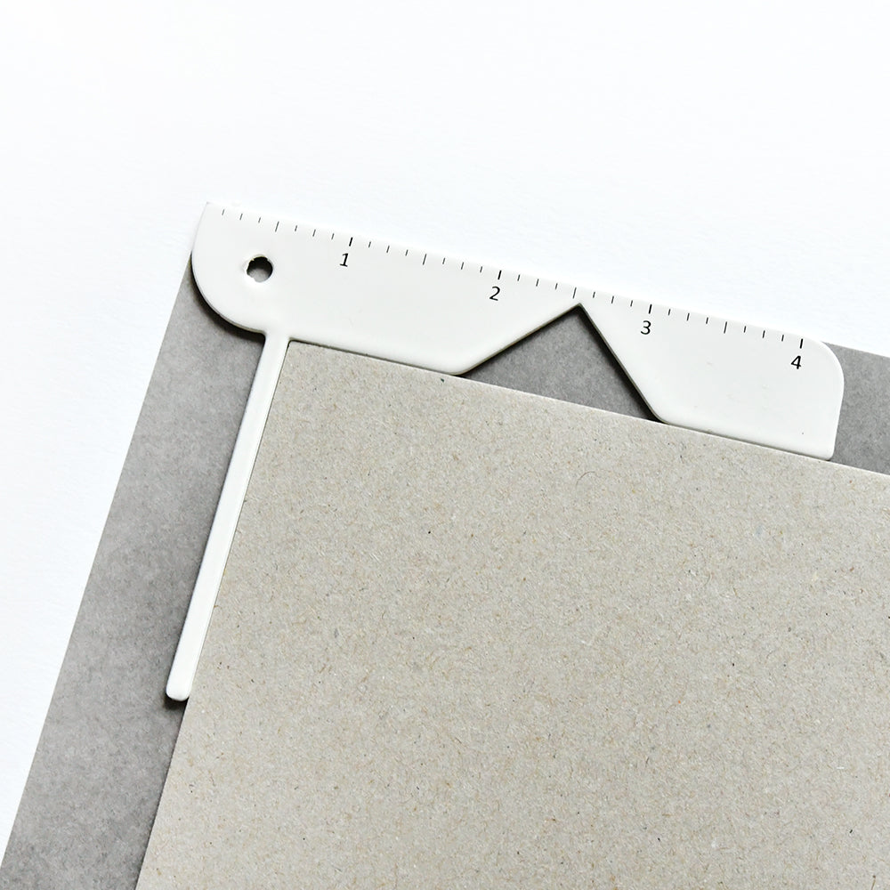 Auf zwei gestapelten grauen und braunen Blättern sitzt ein weißes Vaessen-Messwerkzeug - ideal für Buchhüllen Notizbücher oder das Erstellen von Buchprojekten mit der Bind Easy Buchumschlag-Hilfe.