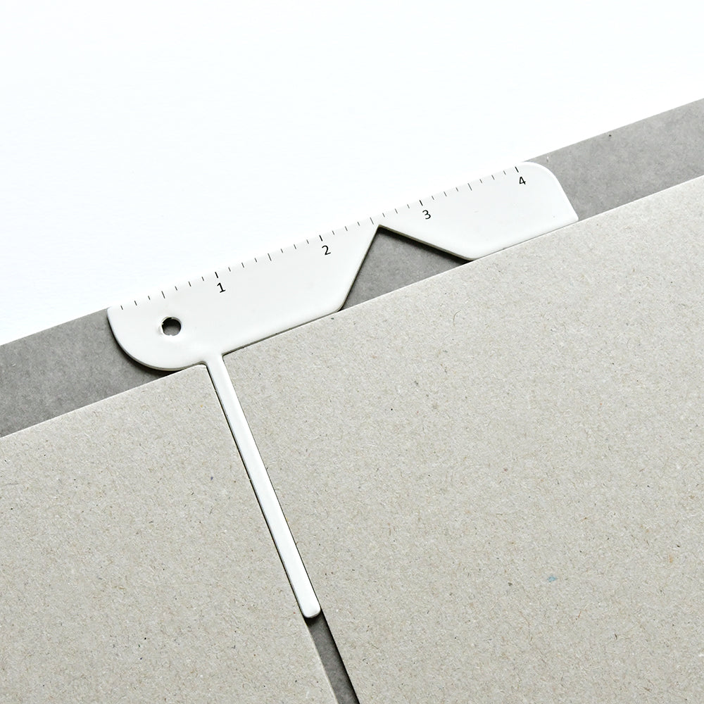 Das weiße Vaessen-Messwerkzeug mit Linealrand richtet zwei Stücke Graukarton präzise aus - ideales Buchbinden-Werkzeug zum Erstellen eines Notizbuch-Umschlags.