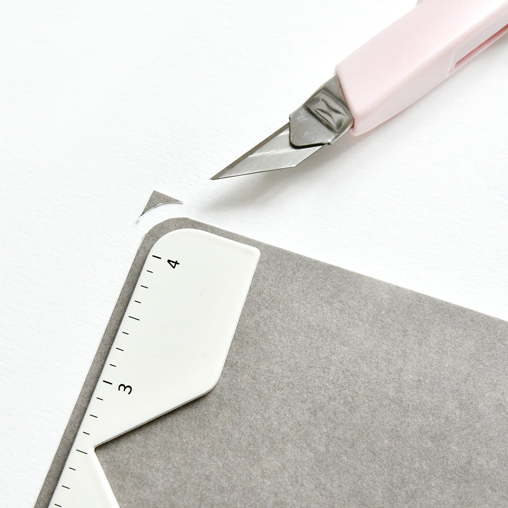 Ein Bastelmesser beschneidet graue Papierecken mit der Vaessen Bind Easy Buchumschlag-Hilfe - eine weiße, abgemessene Eckenführung, ideal zum Buchhüllen basteln und Buchprojekte erstellen.